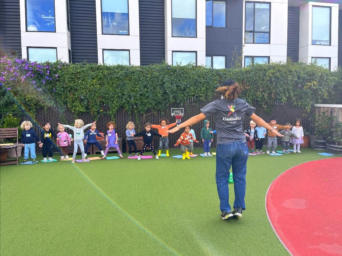 A camp leader with arms outstretched leading preschoolers in a movement game on the Dogpatch campus turf