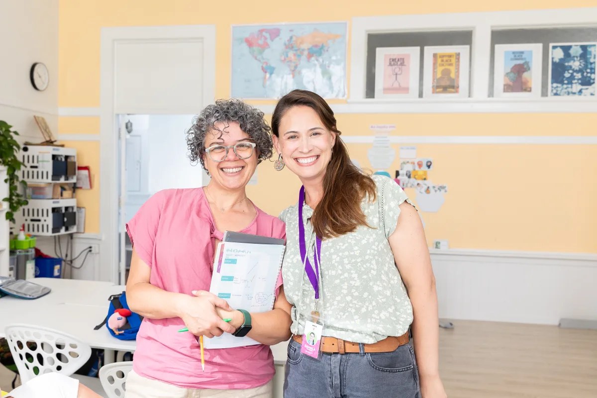 Two La Scuola teachers standing together in a classroom, one holding a booklet